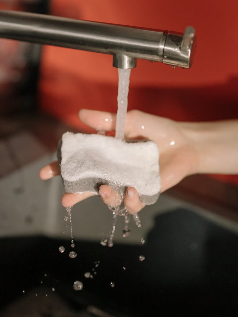 A detailed shot of a hand cleaning a sponge under running water from a tap.