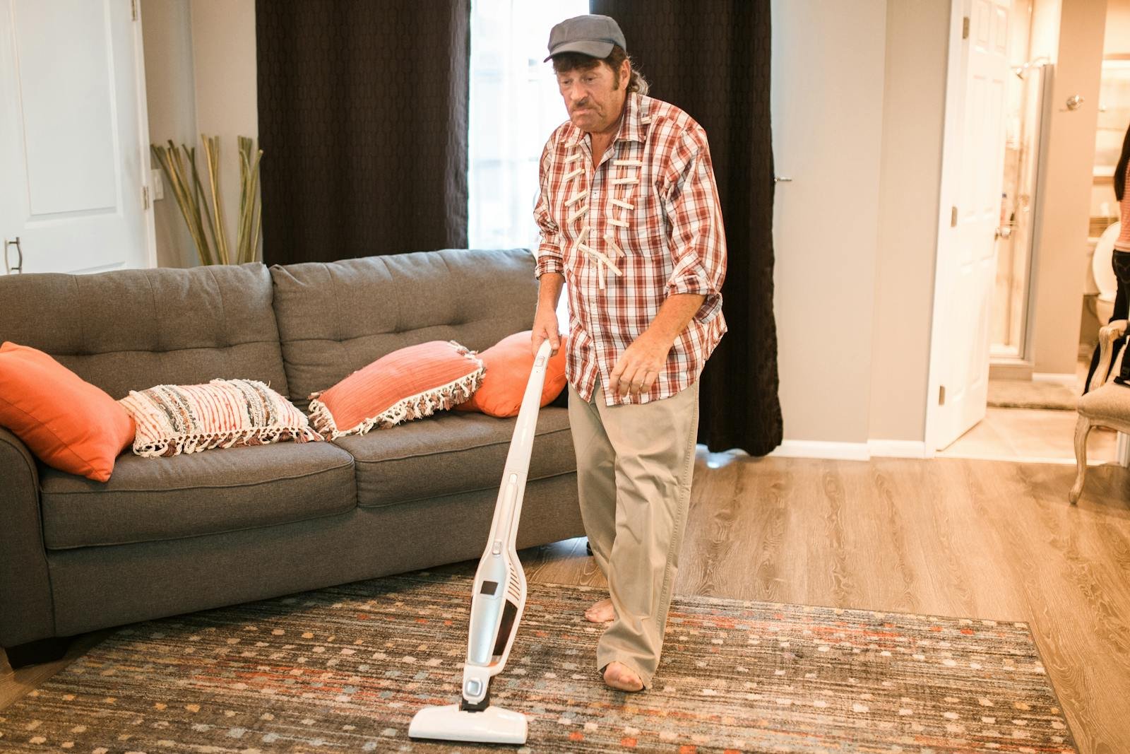 A man cleaning a living room with a vacuum, wearing casual clothes and a checkered shirt.