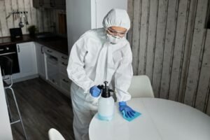 Person wearing PPE cleans a table indoors with sanitizer during pandemic.