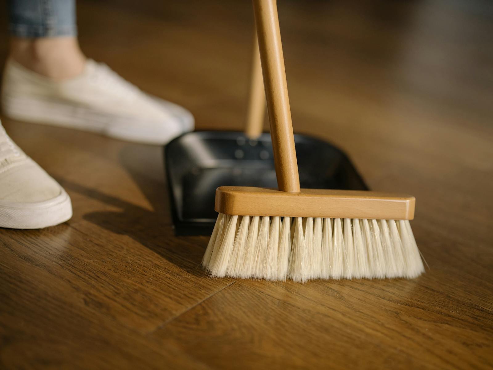 Close-up of a broom and dustpan with white sneakers indoors, representing housekeeping and cleanliness.