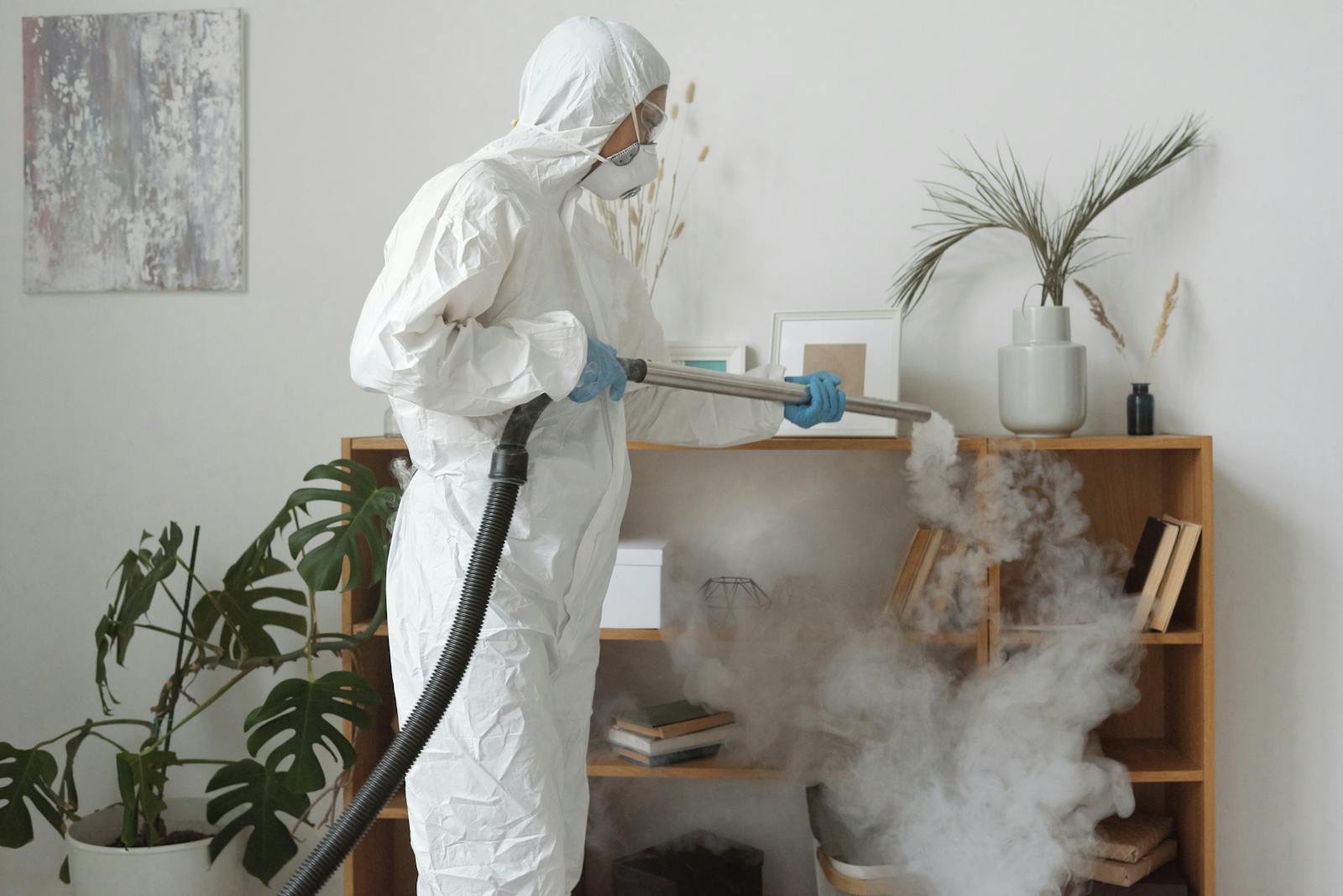 Person using fogging equipment in protective gear to disinfect indoor area with plants and bookshelves.