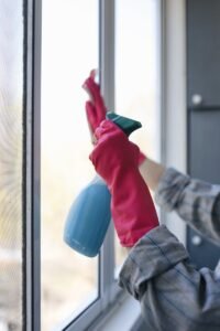 Hands in rubber gloves using a spray bottle to clean a window indoors.