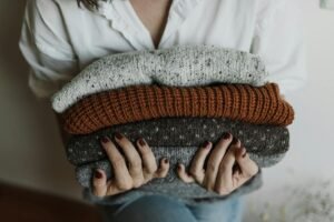 A person holding a stack of folded knitted sweaters in various colors, with a focus on textiles and textures.