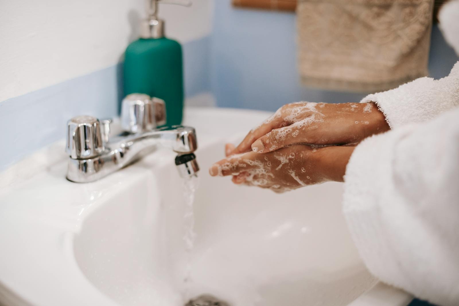 A person in a bathrobe washing hands with soap in a bathroom sink, emphasizing hygiene.