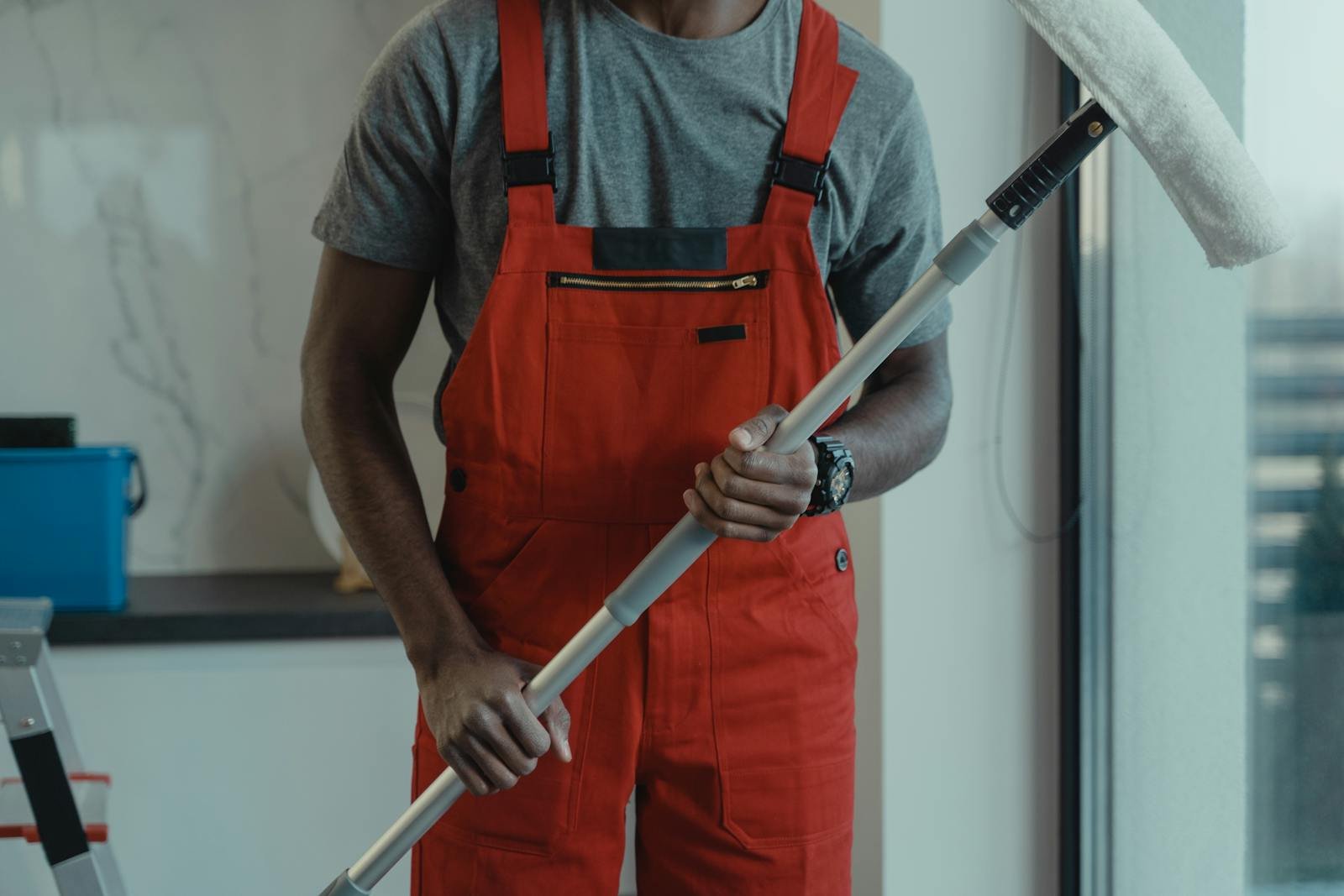 A professional cleaner wearing red overalls is holding a mop while cleaning indoors.