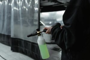 Person in black jacket using power spray tool with cleaning product in an indoor carwash setting.