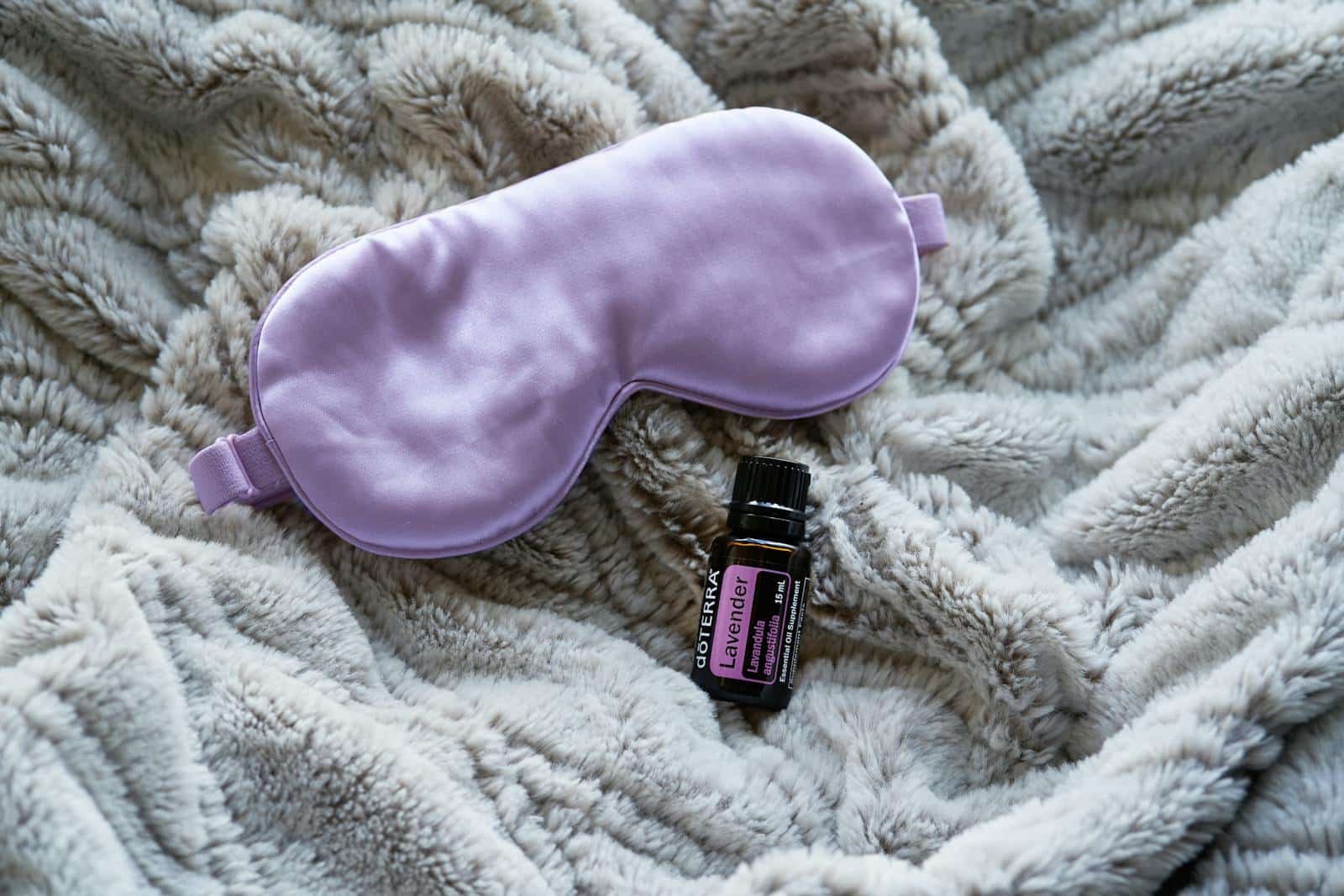 Lavender essential oil alongside a purple sleep mask on a gray blanket promotes relaxation and rest.