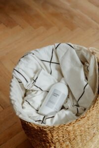 A bottle of laundry liquid rests in a woven basket filled with fabric on a wooden floor.