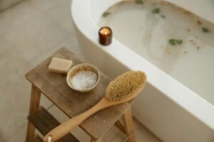 Relaxation bath scene with candle, dry brush, sea salt, and soap on a wooden stool for a serene home spa experience.