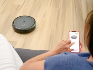 A person uses a smartphone app to control a robotic vacuum on a hardwood floor, showcasing smart home technology.