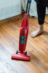 A red vacuum cleaner on a wooden floor with an adult's foot visible, indicating home cleaning.