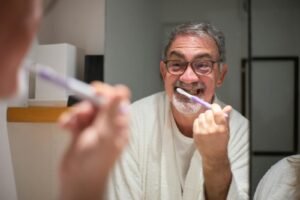 Elderly man in eyeglasses brushes teeth in front of bathroom mirror, smiling warmly.